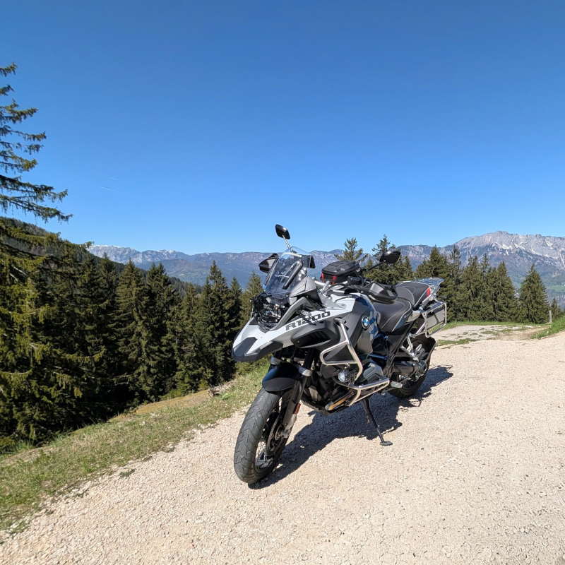 BMW R1200GS Adventure Rallye auf Schotterparkplatz vor bewaldeten Berghängen und Alpengipfeln unter blauem Himmel.