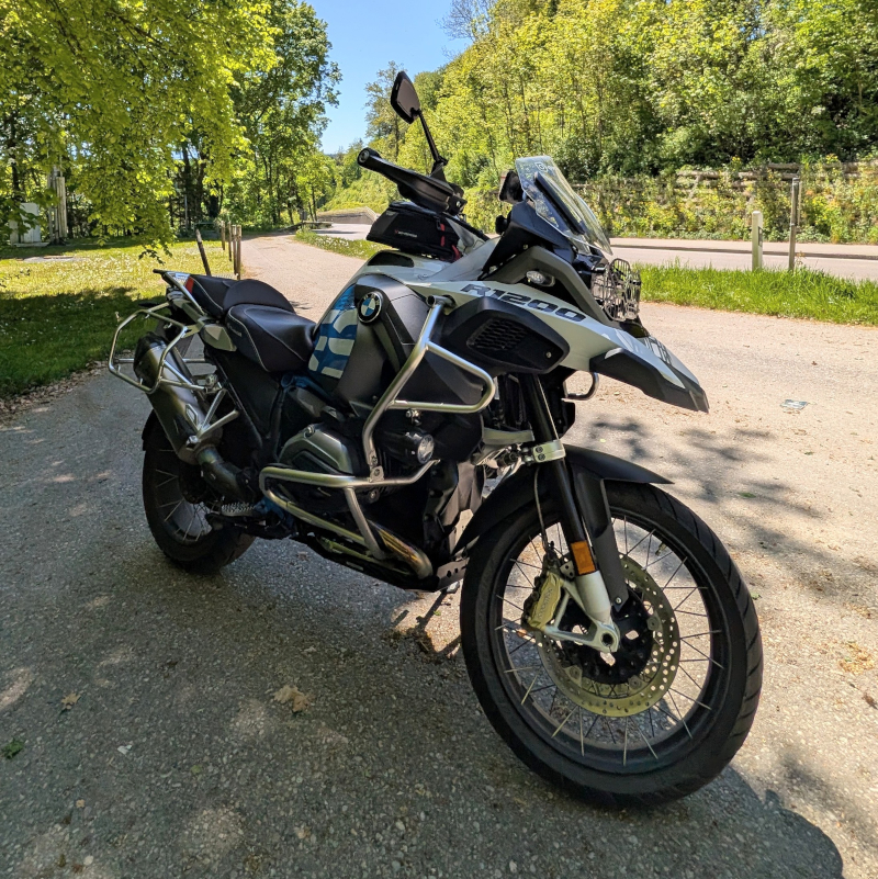 BMW R1200GS Adventure Rallye steht auf sonnigem Waldweg vor einer Kurve. Fokus auf Front und silberne Sturzbügel.