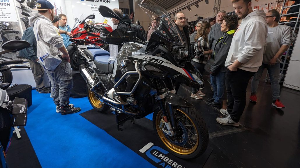 Eine BMW R 1250 GS Adventure mit Carbon-Teilen und goldfarbenen Felgen, umgeben von Besuchern auf einer Messe.