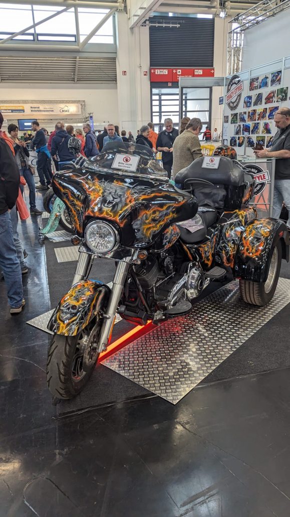 Ein schwarzes Trike mit flammenden Totenkopf-Airbrush-Designs steht auf einer Messe. Rote LED-Beleuchtung unten.