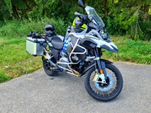BMW R1200 GS in Weiß-Blau mit silbernen Alukoffern, Sturzbügeln und schwarzem Helm auf Asphalt vor grünem Waldrand.