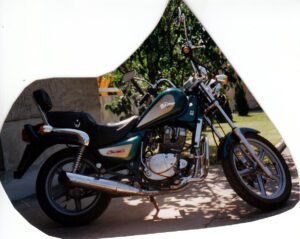 Türkises Chopper-Motorrad mit Chrom-Auspuff, cremefarbenen Akzenten und "Cruise II"-Schriftzug auf sonnigem Stellplatz.