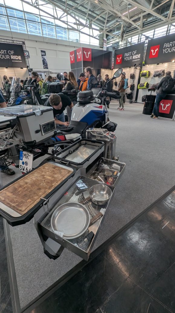 Detailansicht eines BMW-Motorrads auf einem Messe-Stand mit geöffneter, grauer Outdoorküche, Schneidebrett und Kochgeschirr.