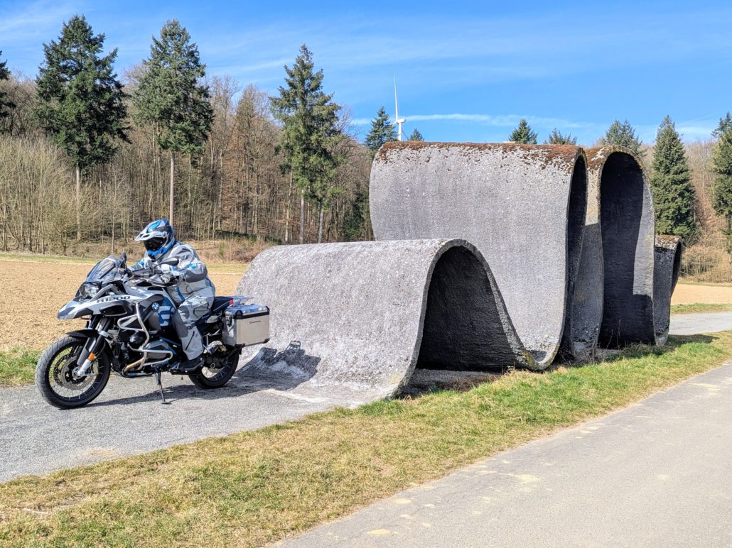 Motorradfahrer auf einer BMW R 1250 GS Adventure neben einer großen, gewellten Beton-Skulptur an einem Feldweg im Wald.
