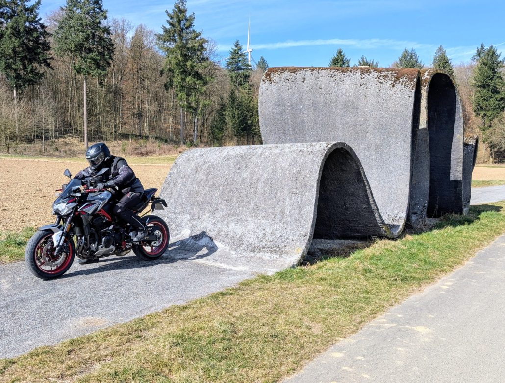 Motorradfahrer fährt auf einer silbernen Kawasaki Z900 mit rotem Rahmen an einer großen grauen, gewellten Beton-Skulptur vorbei.
