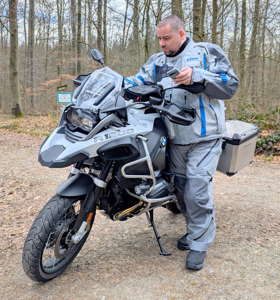 Mann in grauer KLIM-Motorradkleidung lehnt an einer silbernen BMW R 1200 GS mit Seitenkoffer und schaut auf sein Handy.