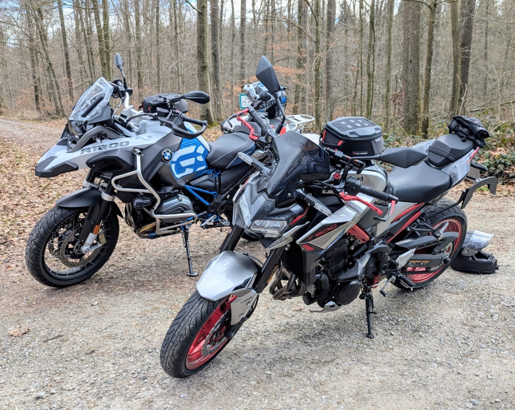 Frontansicht einer silbernen Kawasaki mit rotem Rahmen und Felgen, daneben eine weiße BMW R1200 GS mit Koffern im Wald.