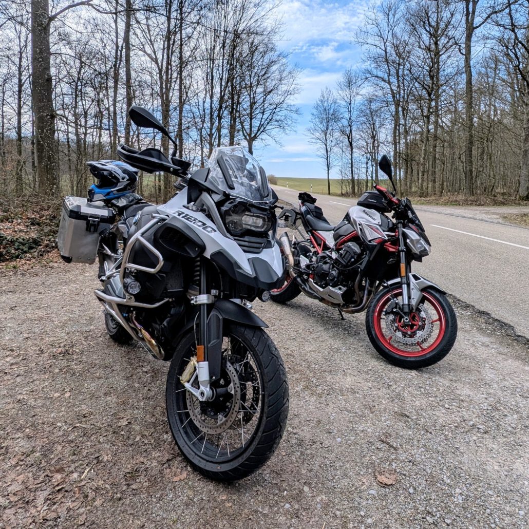 Zwei Motorräder parken am Waldrand: eine weiße BMW GS mit Koffern und eine schwarz-silberne Kawasaki mit roten Felgen.