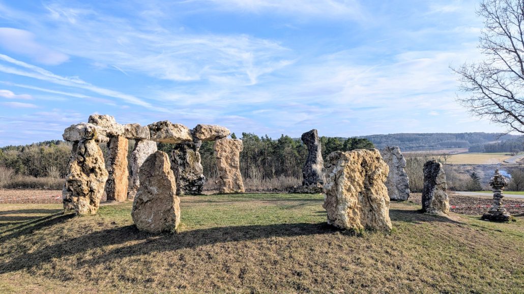 Eine kreisförmige Anordnung von großen, unbearbeiteten Steinen, die an Stonehenge erinnert, auf einem grasbewachsenen Hügel.