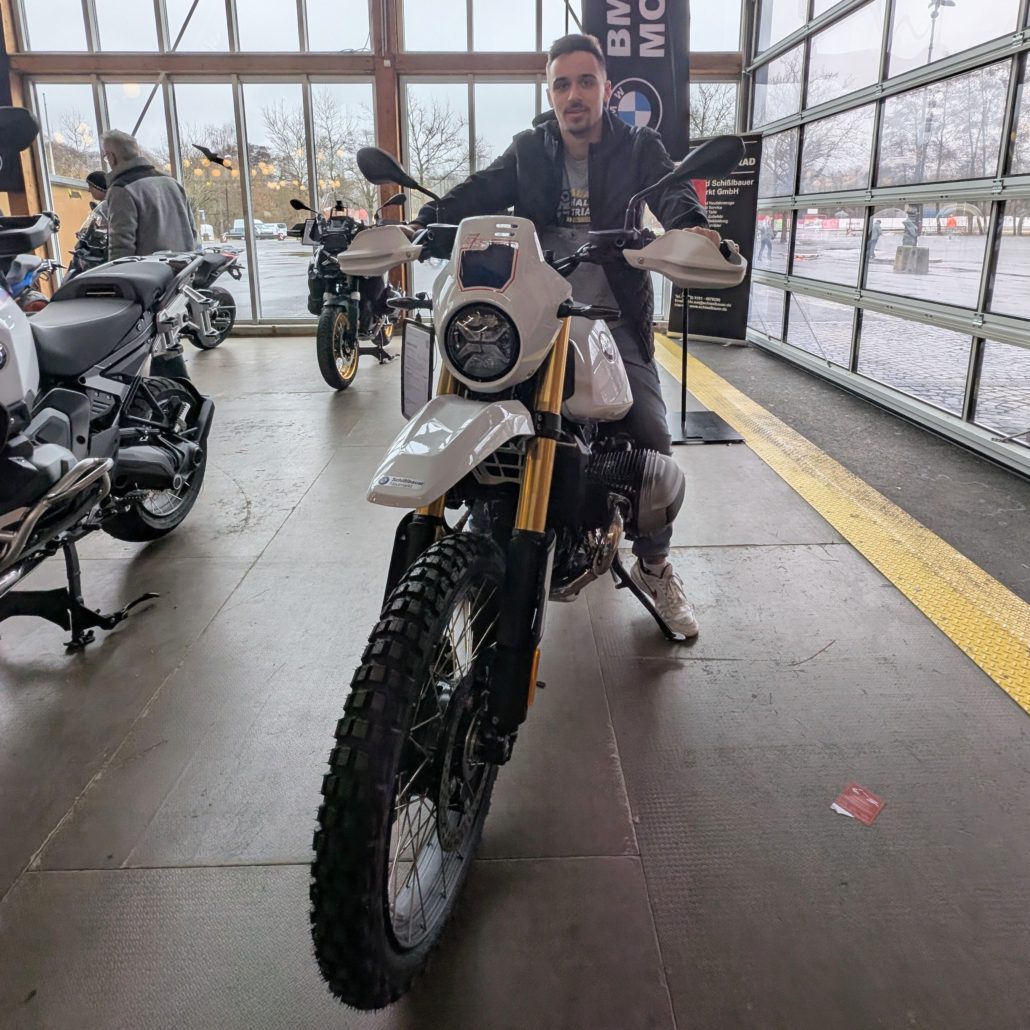 Mann sitzt auf einem weißen BMW-Motorrad mit Stollenreifen und goldenen Federgabeln im Showroom.