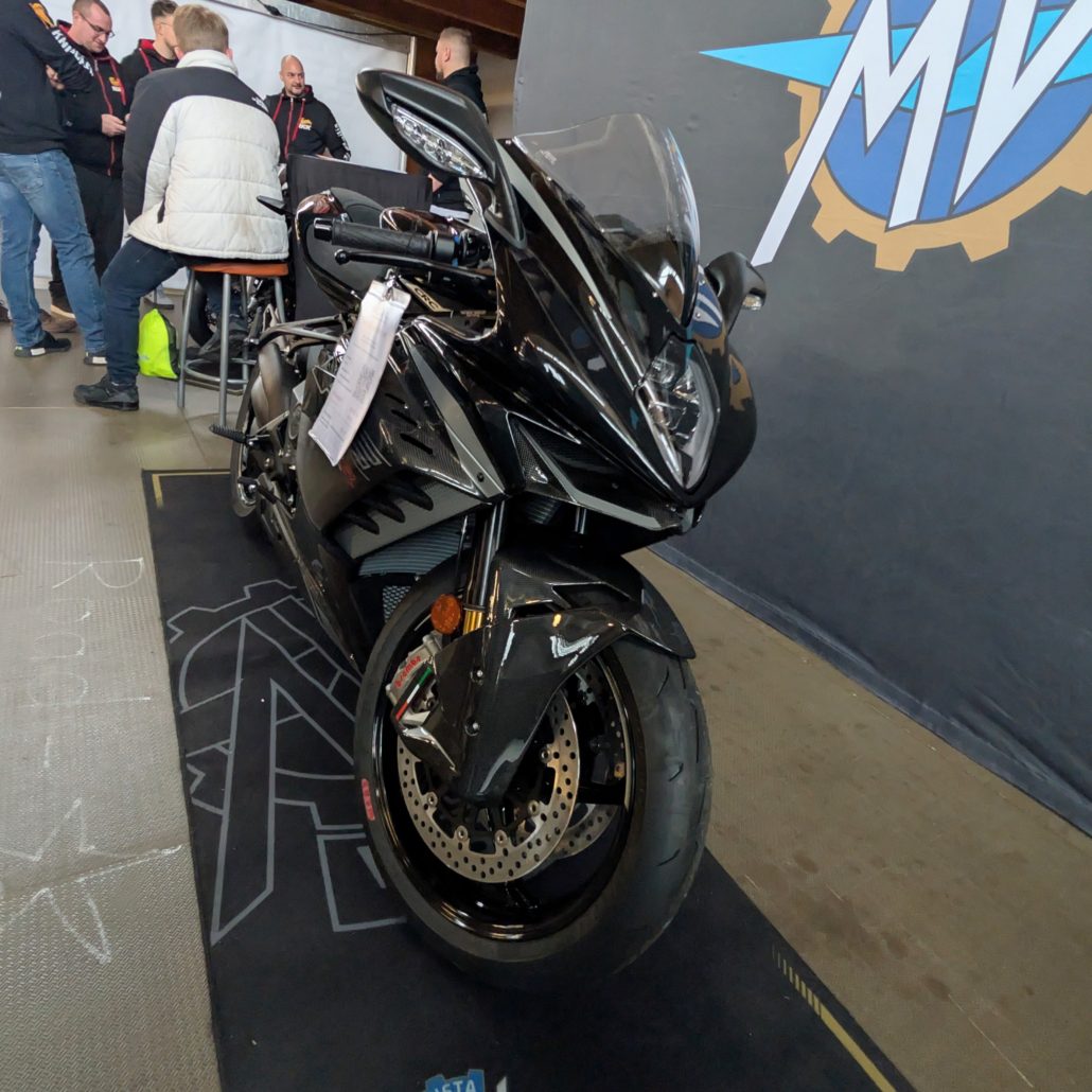 Schwarzes MV Agusta Superbike mit Carbon-Elementen und Brembo-Bremsen auf einem Teppich vor MV-Logo-Hintergrund.