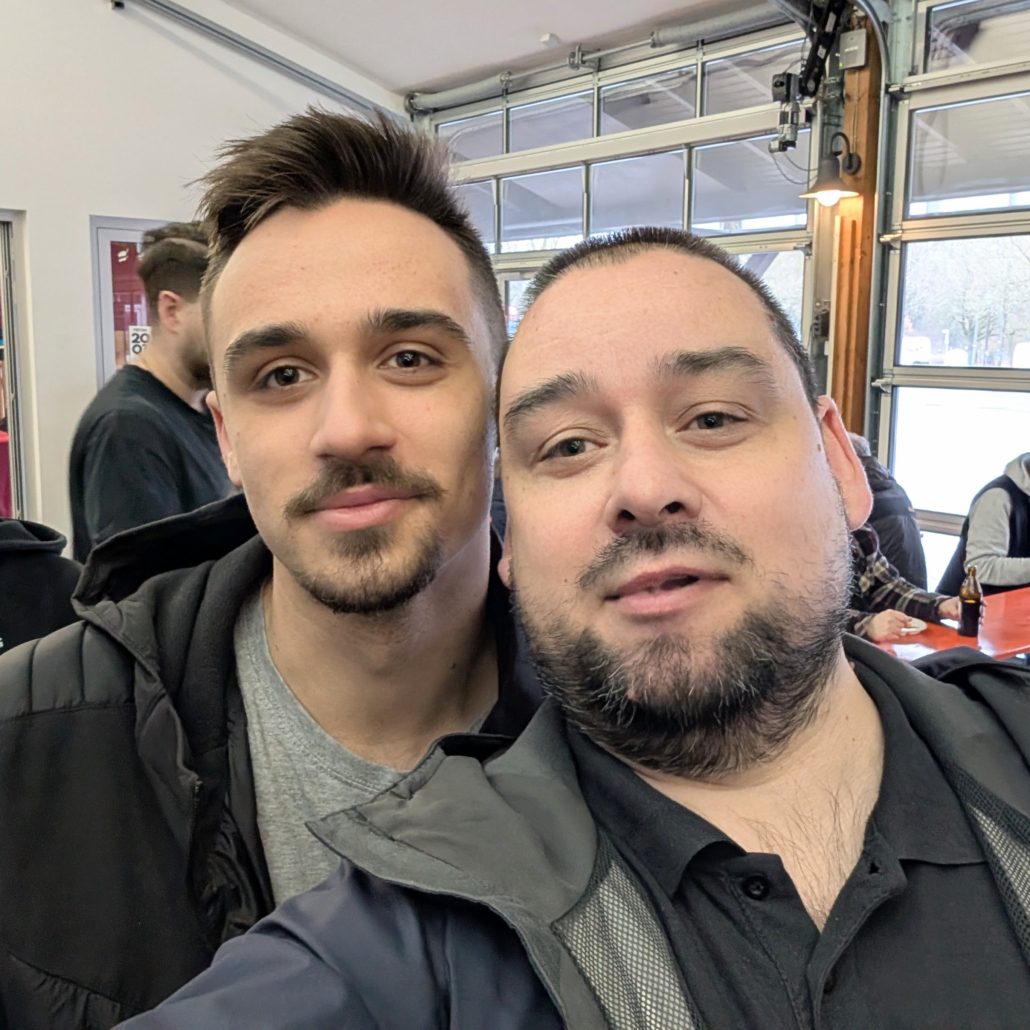 Selfie von zwei Männern mit dunklen Haaren und Bärten in einem hellen Innenraum mit Glasfront im Hintergrund.