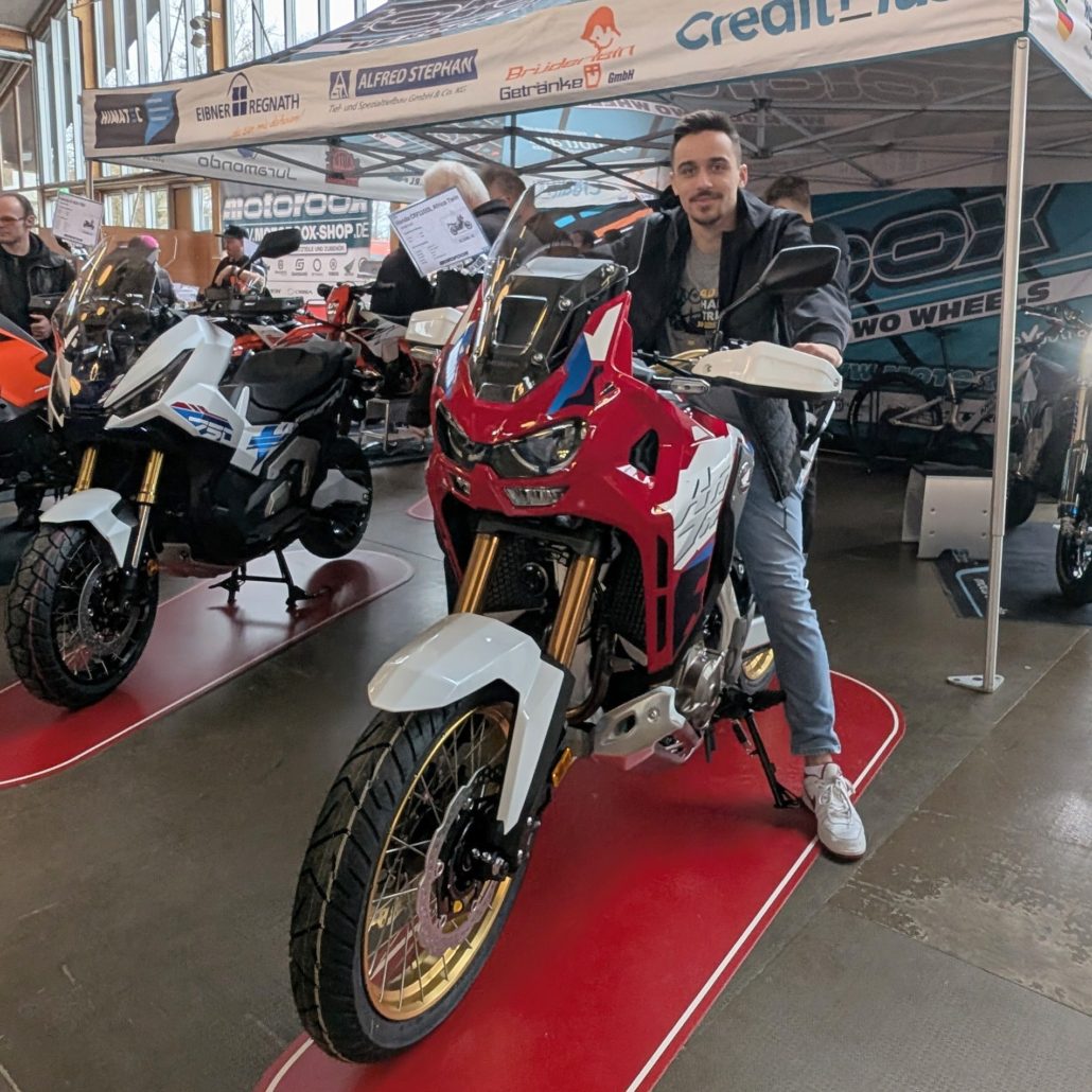 Mann sitzt auf einer rot-weiß-blauen Honda Africa Twin mit goldenen Felgen unter einem Pavillon auf einer Motorradmesse.
