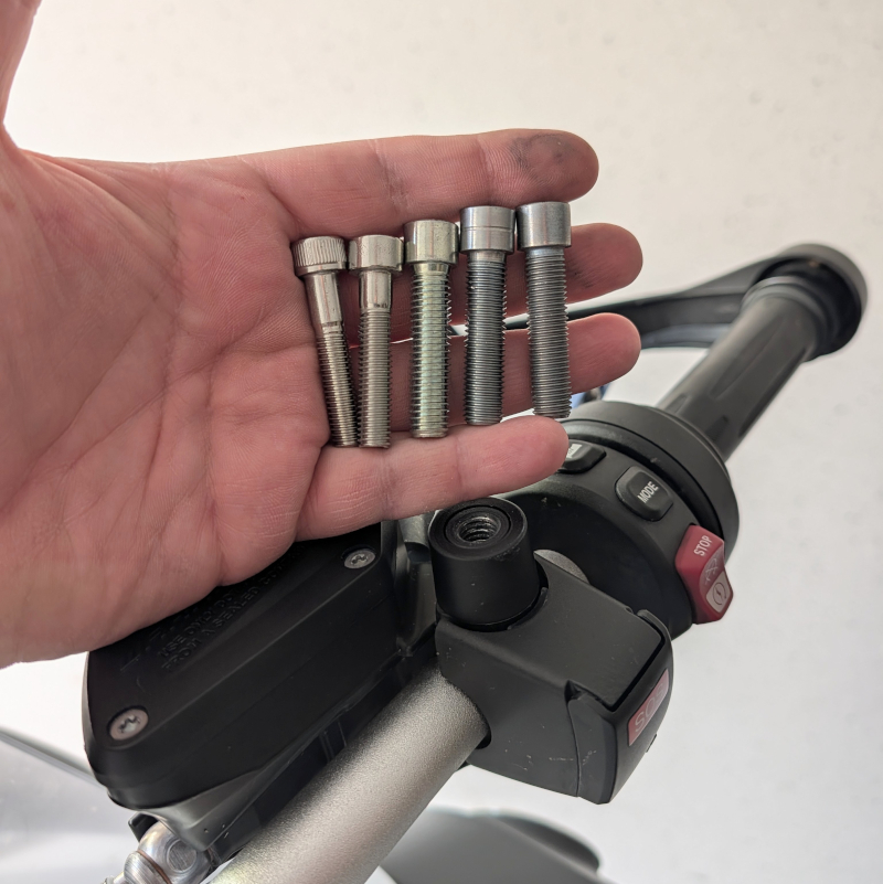 Hand mit fünf silbernen Inbusschrauben aus Metall über einer Motorrad-Armatur mit SOS-Knopf und Bedienelementen.