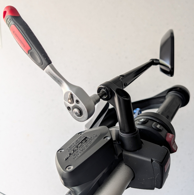 Hebel-Umschaltknarre mit rot-schwarzem Griff wird an schwarzer Metall-Halterung einer Motorrad-Armatur angesetzt.