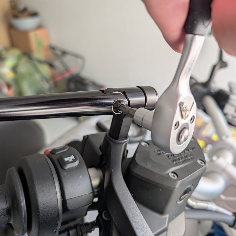 Metall-Knarre zieht Schraube an schwarzem Motorrad-Spiegelarm fest, neben Lenkerarmatur mit Bedienelementen.