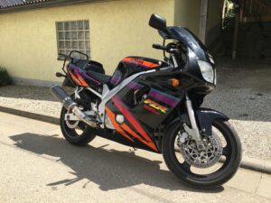 Schwarze Yamaha FZR mit lila-orangem Dekor und "Deltabox Combination"-Schriftzug vor gelbem Haus auf sonniger Straße.