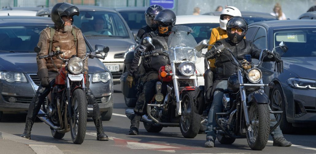Drei Motorradfahrer mit Helmen und Lederkleidung stehen auf ihren Chopper-Motorrädern zwischen stehenden Autos