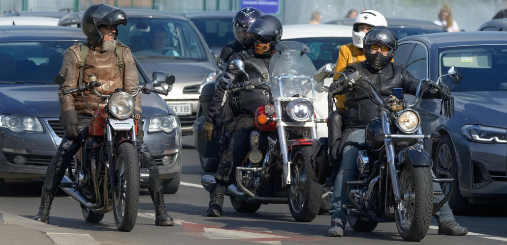 Drei Motorradfahrer mit Helmen und Lederkleidung stehen auf ihren Chopper-Motorrädern zwischen stehenden Autos