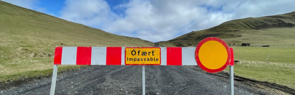 Rot-weiße Straßensperre mit Schild "Ófært Impassable" auf Schotterweg vor grüner Hügellandschaft.