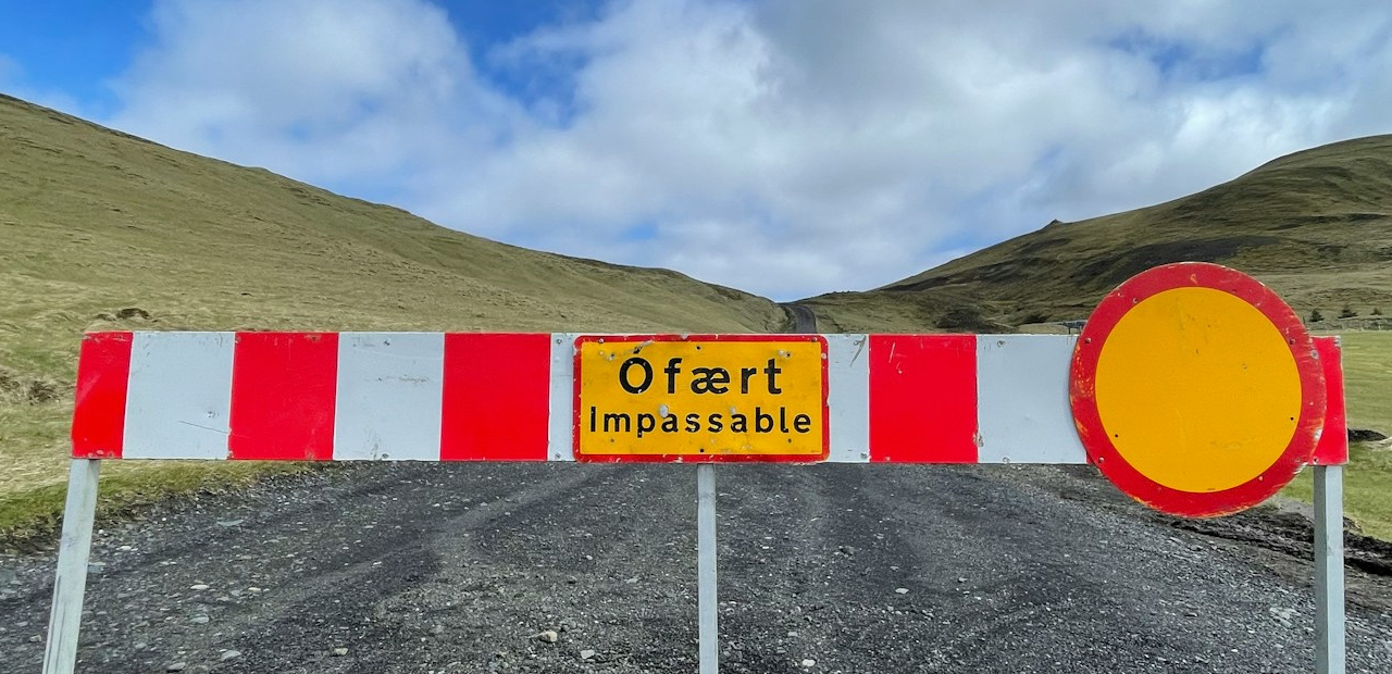 Rot-weiße Straßensperre mit Schild "Ófært Impassable" auf Schotterweg vor grüner Hügellandschaft.