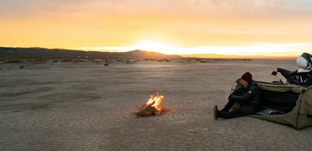 Person sitzt in grünem Zelt vor Lagerfeuer in Wüstenlandschaft bei Sonnenuntergang, schwarzes Motorrad steht daneben.