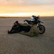 Person sitzt in grünem Zelt vor Lagerfeuer in Wüstenlandschaft bei Sonnenuntergang, schwarzes Motorrad steht daneben.