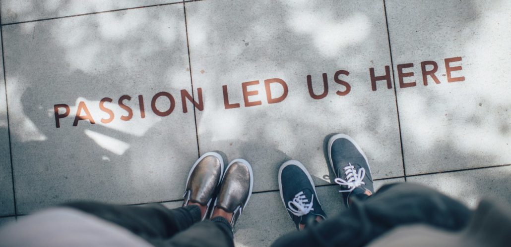 Zwei Paar Füße auf einem hellgrauen Boden; darüber steht in großen braunen Buchstaben: „PASSION LED US HERE“