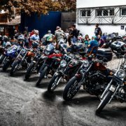 Lange Reihe geparkter Motorräder am Straßenrand und stehende Biker-Gruppen vor einem Gebäude bei einem Treffen.