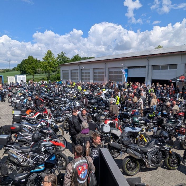Großes Motorradtreffen mit zahlreichen geparkten Maschinen und Bikern in Schutzkleidung vor einer Halle bei Sonnenschein.