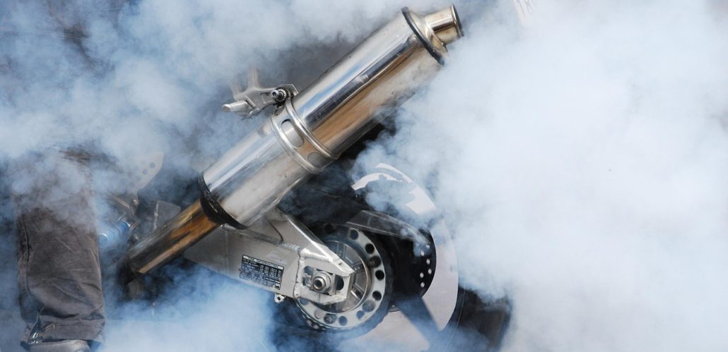 Ein glänzender, verchromter Motorrad-Auspuff, umgeben von dichtem weißem Rauch aus dem Endschalldämpfer und den durchdrehenden Reifen